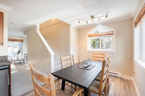 Un coin repas avec une table en bois foncé, six chaises en bois, un escalier et une fenêtre avec des stores.