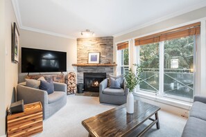 A living room with a fireplace, a flat-screen TV, and a large window with a view of trees. - Private Bike Storage | Free parking | Mountain Views | Window AC (Whistler)