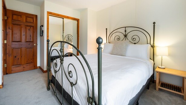 Quarto com porta de madeira, estrutura de cama de metal, mesa de cabeceira com abajur e espelho na parede.