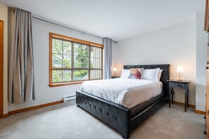 A bedroom with a large window, a bed, a bedside table, and a nightstand. - Spacious townhome | Private garage & hot tub | BBQ (Whistler)