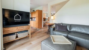 Uma sala de estar moderna com televisão de tela plana, um sofá de couro cinza, uma mesa de centro de madeira e uma área de cozinha ao fundo.