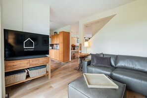 Ein modernes Wohnzimmer mit Flachbildfernseher, grauem Ledersofa, Couchtisch aus Holz und einer Küche im Hintergrund.