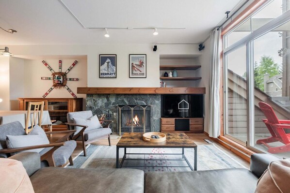 A modern living room with a stone fireplace, comfortable seating, and a large window. - Spacious townhome | Private garage & hot tub | BBQ (Whistler)