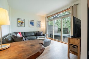 Un salon moderne avec un canapé, une table basse et un téléviseur posé sur un meuble en bois. De grandes fenêtres offrent une vue sur la verdure environnante.