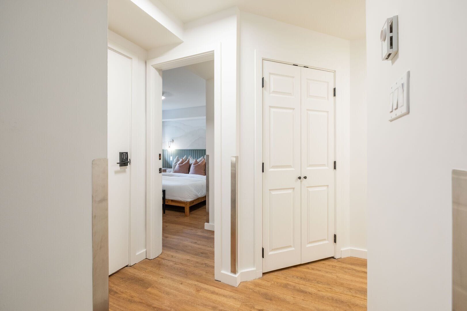 Ein Flur, der zu einem Schlafzimmer führt, mit einem Bett, einer Tür und einem Lichtschalter an der Wand.