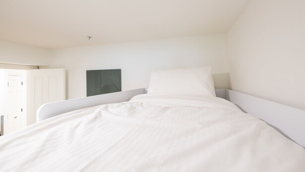 Una cama individual con ropa de cama blanca en una habitación con paredes y una puerta blancos.