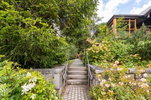 A stone staircase leading to a building with a wooden facade, surrounded by lush greenery and flowering plants. - Private hot tub | Pets | BBQ | Location! (Whistler)