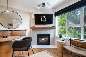 A modern living room with a fireplace, a television mounted above it, and a large window providing a view of greenery outside. - Private hot tub | Pets | BBQ | Location! (Whistler)
