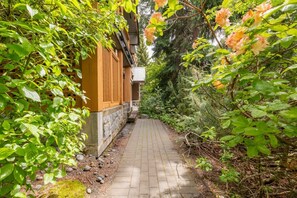 A narrow brick pathway leading to a building with wooden shutters and a stone base. - Private hot tub | Pets | BBQ | Location! (Whistler)