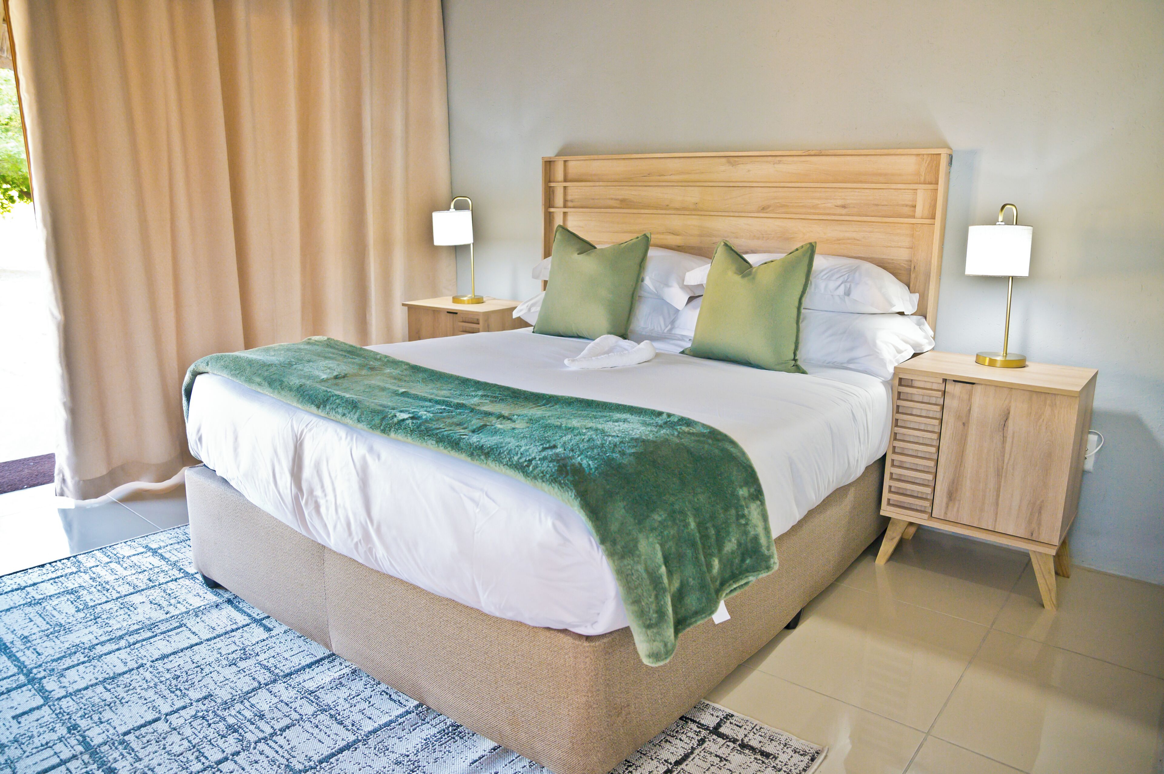 A bed with a wooden headboard, two bedside tables with lamps, a green throw blanket, and a patterned rug.