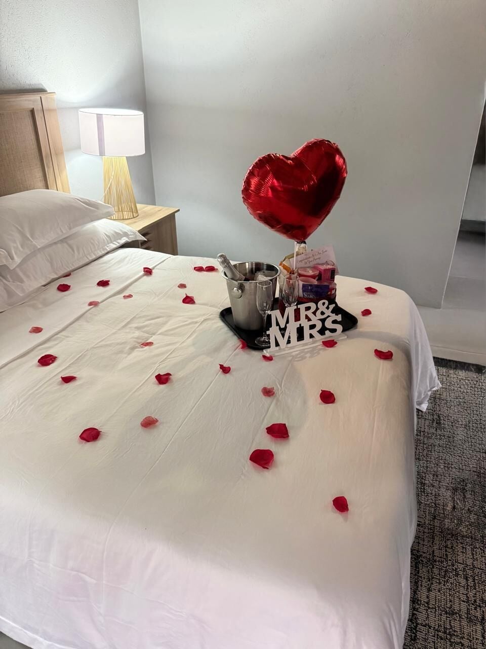 Un lit recouvert de pétales de rose, un ballon en forme de cœur, un seau à champagne et une pancarte sur laquelle on peut lire « M. et Mme ».