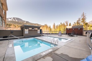 A rooftop pool with a view of mountains and trees, surrounded by buildings and outdoor seating. - Stunning ski-in/out | AC | Complex pool & hot tub (Whistler)