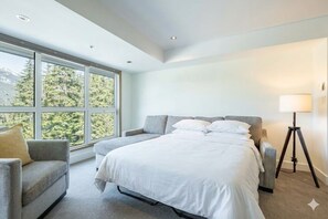 Habitación de hotel moderna con una cama grande, un sofá, una lámpara de pie y vistas a un bosque a través de amplios ventanales.