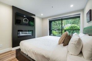 A modern bedroom with a large bed, a TV mounted on the wall, and a window with a view of greenery. - Walk to Blackcomb | Complex hot tub | BBQ (Whistler)