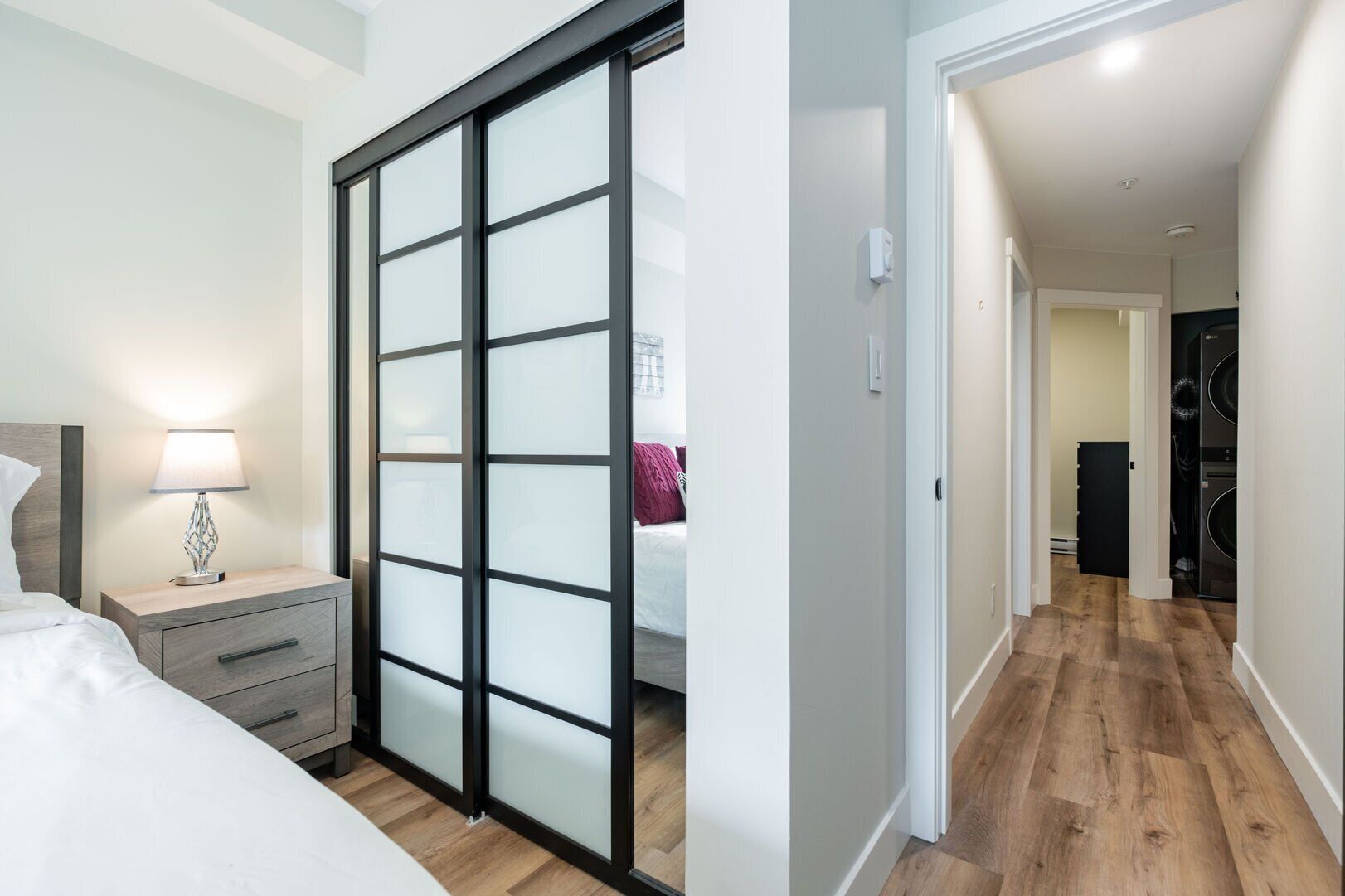 A bedroom with a bed, nightstand, lamp, and a sliding glass door leading to a hallway with a washing machine.