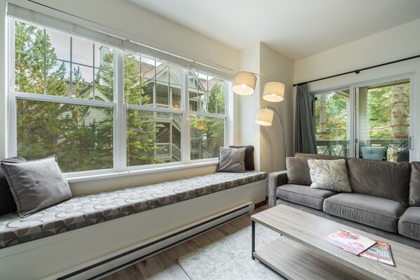 Ein modernes Wohnzimmer mit großen Fenstern, einer integrierten Sitzbank, einem grauen Sofa und einem Couchtisch mit Zeitschriften.