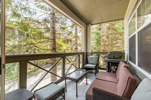 Eine überdachte Veranda mit Korbmöbeln, einem Grill und Blick auf Bäume.