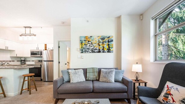 A modern living room with a grey sofa, a coffee table, and a chair with a festive pillow. A kitchen is visible in the background. - Beautiful Condo | Shared hot tub | BBQ | Wifi (Whistler)