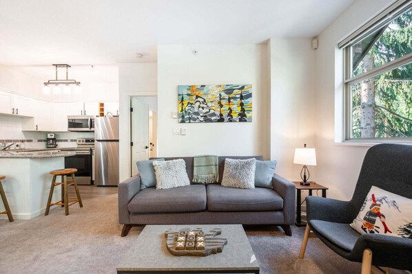 A modern living room with a grey sofa, a coffee table, and a chair with a festive pillow. A kitchen is visible in the background. - Beautiful Condo | Shared hot tub | BBQ | Wifi (Whistler)