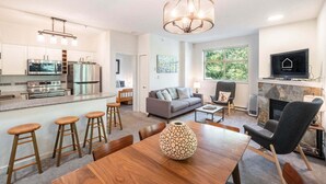 A modern kitchen with stainless steel appliances, a dining area with wooden stools, a grey sofa, and a fireplace. - Beautiful Condo | Shared hot tub | BBQ | Wifi (Whistler)