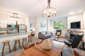 A modern kitchen with stainless steel appliances, a dining area with wooden stools, a grey sofa, and a fireplace. - Beautiful Condo | Shared hot tub | BBQ | Wifi (Whistler)