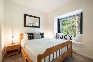Une chambre avec un lit en bois, une table de chevet, un banc près de la fenêtre agrémenté de coussins et un miroir au mur.