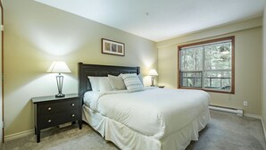 A bedroom with a bed, two bedside tables with lamps, a window with a view of trees, and a wall-mounted picture. - Ski-in/out apartment | Complex pool, hot tub & gym (Whistler)