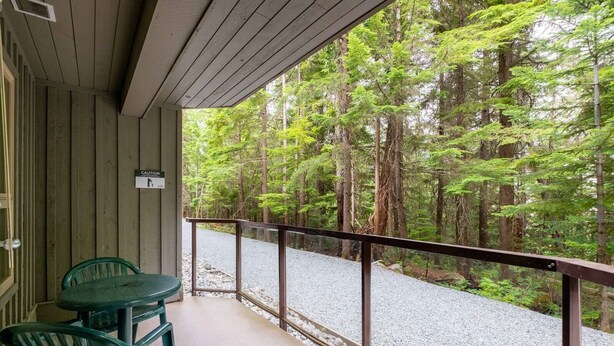 A covered porch with a table and chairs, overlooking a gravel path and dense forest. - Ski-in/out apartment | Complex pool, hot tub & gym (Whistler)