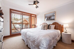 A bedroom with a large bed, a wooden headboard, a ceiling fan, a bedside table with a lamp, and a large sliding glass door leading to an outdoor area with a deck and chairs. - Townhome | Private hot tub | BBQ | A/C | Pets (Whistler)