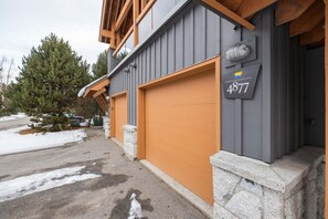 A building with a grey exterior and a wooden garage door. A mailbox with the number 4877 is attached to the wall. - Townhome | Private hot tub | BBQ | A/C | Pets (Whistler)