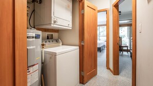 A laundry room with a front-loading washer and dryer, wooden doors, and a view into a bedroom with a bed and a chair. - Great location | Complex hot tub | BBQ | Smart TVs (Whistler)