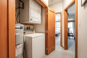 Ein Waschraum mit einer Waschmaschine und einem Trockner, Holztüren und Blick in ein Schlafzimmer mit Bett und Sessel.