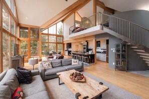 A spacious living area with a large sofa, a coffee table, and a dining table. There is a kitchen area with modern appliances and a staircase leading to an upper level. - Luxury Home | Private hot tub | Pets | BBQ | AC (Whistler)