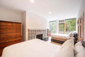 A bedroom with a large bed, a dresser, a fireplace, and a view of trees through large windows. - Luxury Home | Private hot tub | Pets | BBQ | AC (Whistler)