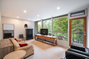 Ein modernes Wohnzimmer mit Kamin, Fernseher, einem Holztisch und großen Fenstern mit Blick auf einen Wald.
