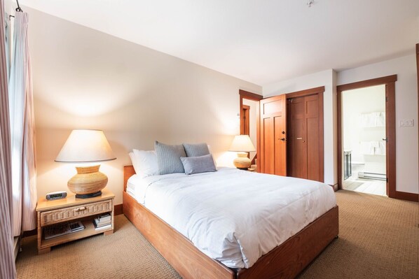 A neatly arranged bedroom with a large bed, two bedside tables with lamps, a wooden wardrobe, and a door leading to a bathroom. - Townhome | Smart TVs | Pets | BBQ | Blueberry Hill (Whistler)