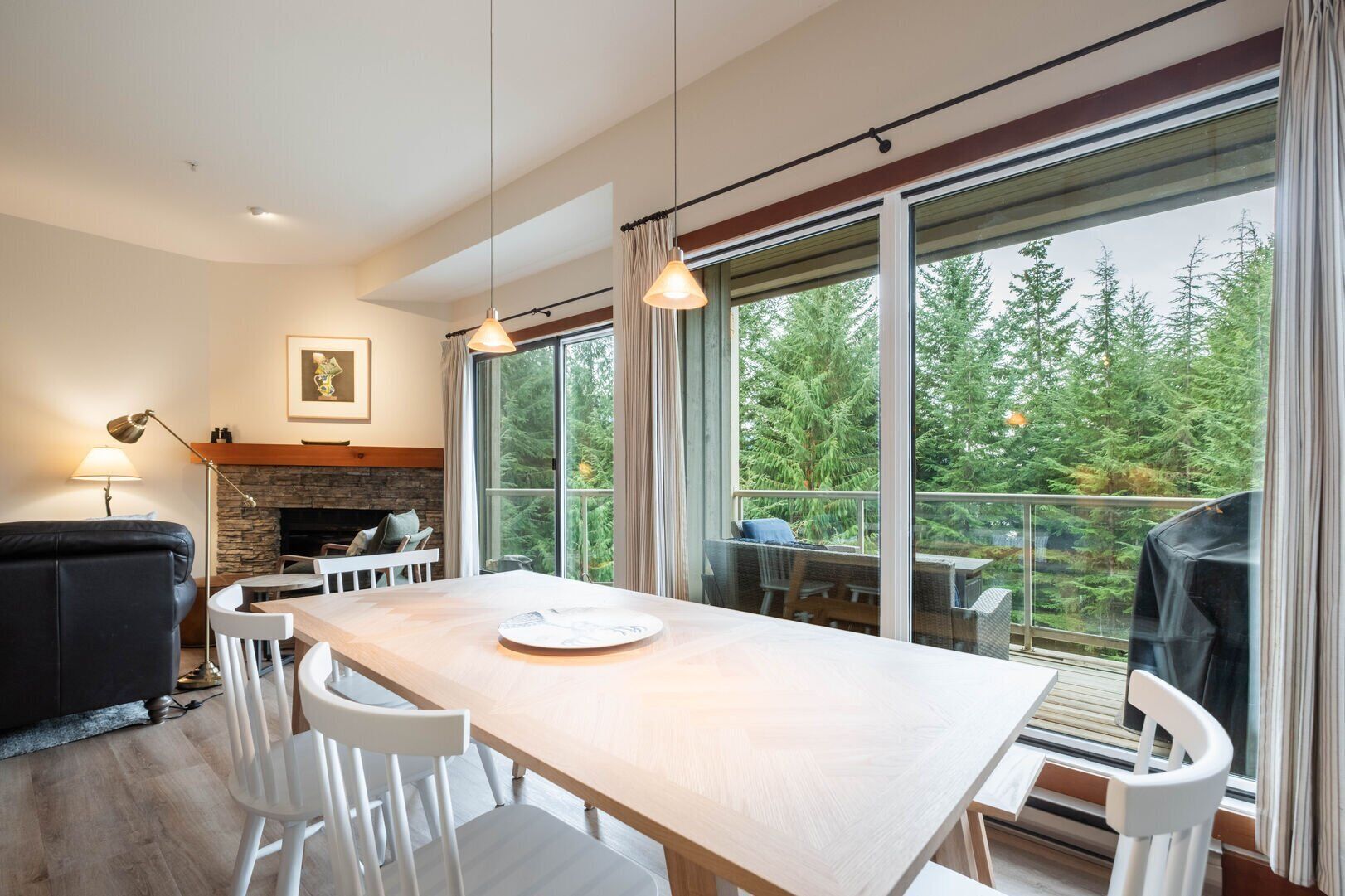 A modern living room with a fireplace, a dining table, and large windows offering a view of trees.
