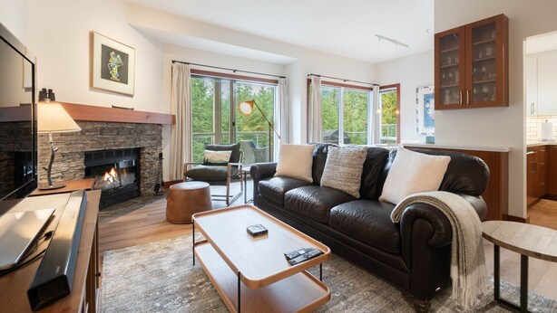A modern living room with a stone fireplace, leather sofa, and a dining area with a wooden table and bench. - Townhome | Smart TVs | Pets | BBQ | Blueberry Hill (Whistler)