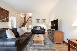 A living room with a black leather sofa, a wooden coffee table, a television, and a staircase leading to an upper floor. - Townhome | Smart TVs | Pets | BBQ | Blueberry Hill (Whistler)