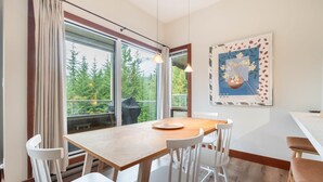 A dining area with a wooden table and white chairs, large windows with a view of trees, and a framed artwork on the wall. - Townhome | Smart TVs | Pets | BBQ | Blueberry Hill (Whistler)