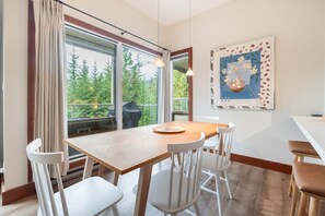 Ein Essbereich mit einem Holztisch und weißen Stühlen, großen Fenstern mit Blick auf Bäume und einem gerahmten Kunstwerk an der Wand.