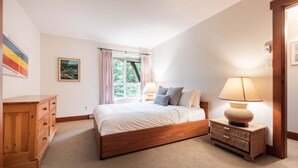 A bedroom with a wooden bed, a nightstand with a lamp, a dresser, a window with curtains, and a framed picture on the wall. - Townhome | Smart TVs | Pets | BBQ | Blueberry Hill (Whistler)