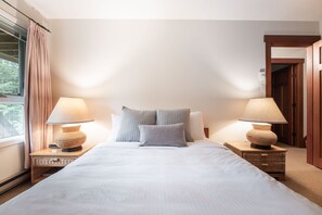 A neatly made bed with two pillows, two bedside tables with lamps, and a view of a door and a window with curtains. - Townhome | Smart TVs | Pets | BBQ | Blueberry Hill (Whistler)