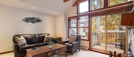 A living room with a leather couch, a wooden coffee table, a grey armchair, and a large sliding glass door leading to a balcony.