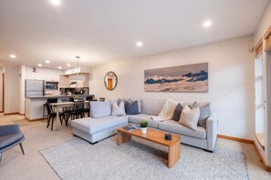 A modern living room with a sectional sofa, a dining area with chairs, a kitchen with appliances, and a large window with a mountain view. - Townhome BBQ | Smart TV | Private Garage | Wifi (Whistler)