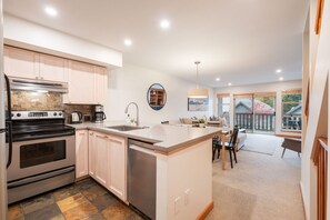 A modern kitchen with a central island, built-in oven, and dishwasher. There is a dining area with a table and chairs, and a view of the outside through large windows. - Townhome BBQ | Smart TV | Private Garage | Wifi (Whistler)