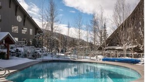 En swimmingpool med ett trädäck, omgiven av snötäckta träd och en byggnad med stora fönster.