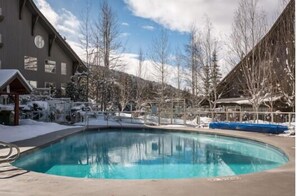A swimming pool with a wooden deck, surrounded by snow-covered trees and a building with large windows. - Condo | Ski-in/ski-out | Shared pool & hot tub (Whistler)
