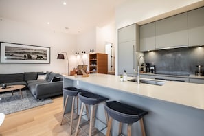 Eine moderne Wohnung mit einem grauen Sofa, einem kleinen Couchtisch, einer Küche mit grauen Schränken und Edelstahlgeräten sowie einem großen Fenster.