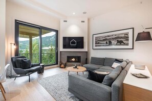 A modern living room with a fireplace, a sofa set, a coffee table, and a large window offering a view of a forested area. - Townhome | Pets | BBQ | Free park | SmartTV | Wifi (Whistler)